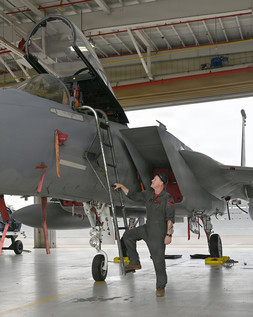 A military pilot or aircrew member in an olive-green flight suit is conducting a pre-flight or post-flight inspection of a gray fighter jet, identified by visible markings as an F-15 Eagle. The aircraft is positioned inside or at the entrance of a military hangar, with the cockpit canopy open and an access ladder deployed. The individual is looking upward toward the cockpit area while standing on the ladder steps.
