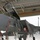104th Fighter Wing Celebrates Flight Surgeon's Fifteen Years of Service with Fini Flight A military pilot or aircrew member in an olive-green flight suit is conducting a pre-flight or post-flight inspection of a gray fighter jet, identified by visible markings as an F-15 Eagle. The aircraft is positioned inside or at the entrance of a military hangar, with the cockpit canopy open and an access ladder deployed. The individual is looking upward toward the cockpit area while standing on the ladder steps.