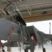 A military pilot or aircrew member in an olive-green flight suit is conducting a pre-flight or post-flight inspection of a gray fighter jet, identified by visible markings as an F-15 Eagle. The aircraft is positioned inside or at the entrance of a military hangar, with the cockpit canopy open and an access ladder deployed. The individual is looking upward toward the cockpit area while standing on the ladder steps.