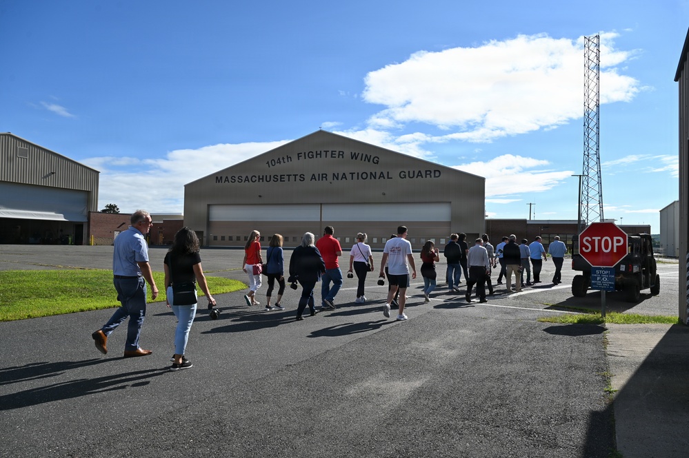 A group of civilians is walking toward a large military hangar belonging to the 104th Fighter Wing, Massachusetts Air National Guard. The scene appears to be an organized tour or public visit to the air base facility. The hangar is prominently labeled and serves as the focal point of the image.