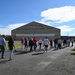 A group of civilians is walking toward a large military hangar belonging to the 104th Fighter Wing, Massachusetts Air National Guard. The scene appears to be an organized tour or public visit to the air base facility. The hangar is prominently labeled and serves as the focal point of the image.