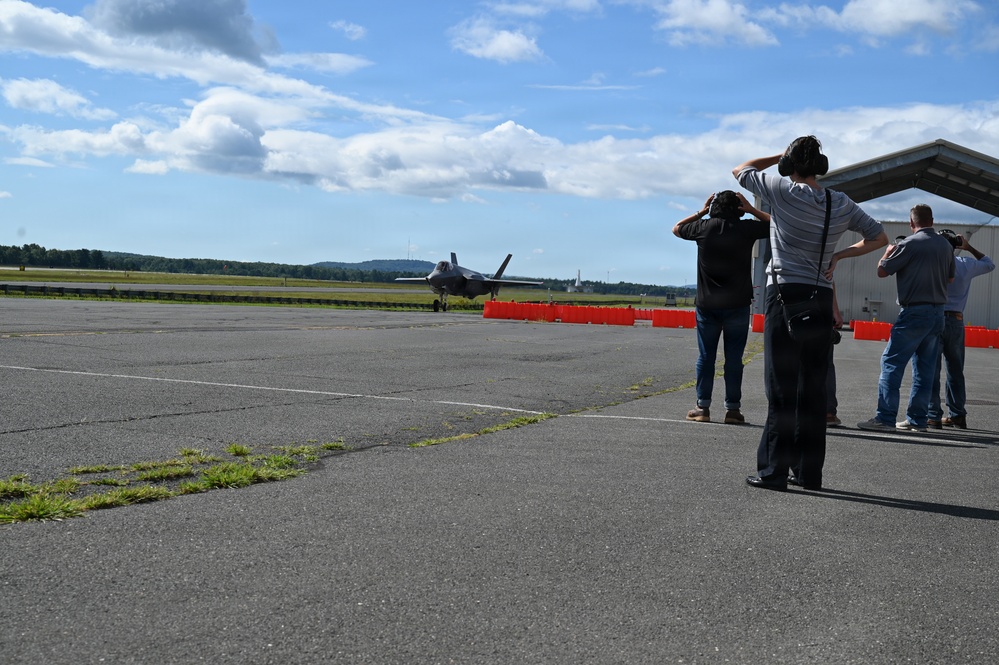 A group of observers and photographers watch a military jet aircraft, appearing to be an F-35, on a runway or taxiway at an airfield. Several people are shielding their eyes or raising cameras to document the aircraft's movement. Orange traffic cones or barriers are visible in the background delineating a restricted area.