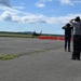 A group of observers and photographers watch a military jet aircraft, appearing to be an F-35, on a runway or taxiway at an airfield. Several people are shielding their eyes or raising cameras to document the aircraft's movement. Orange traffic cones or barriers are visible in the background delineating a restricted area.