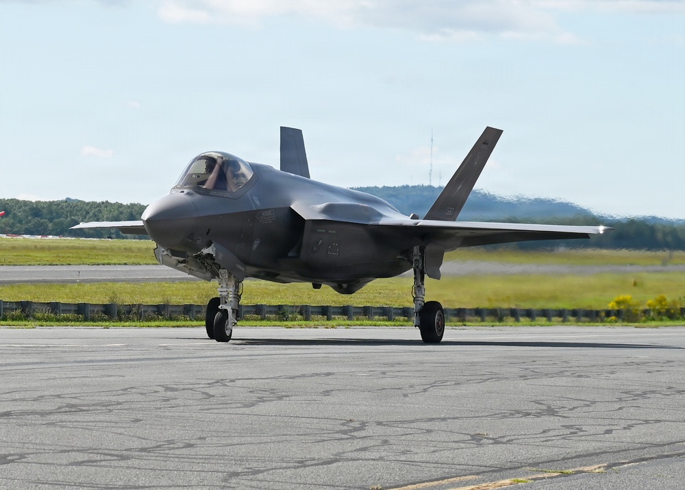 A single-seat stealth fighter jet, identifiable as an F-35 Lightning II, is taxiing on an airfield runway or taxiway. The aircraft is painted in a dark gray low-observable finish and has its landing gear fully deployed. The setting appears to be a military or joint-use airfield surrounded by green terrain and hills in the background.
