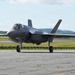 A single-seat stealth fighter jet, identifiable as an F-35 Lightning II, is taxiing on an airfield runway or taxiway. The aircraft is painted in a dark gray low-observable finish and has its landing gear fully deployed. The setting appears to be a military or joint-use airfield surrounded by green terrain and hills in the background.