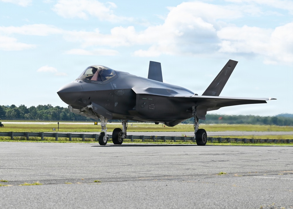A single-engine stealth multirole fighter aircraft, consistent with an F-35 Lightning II, is taxiing on a paved runway or taxiway. The aircraft is photographed in daylight under partly cloudy skies, with green treelines and grass visible in the background. A pilot is visible in the cockpit, and the landing gear is fully deployed.