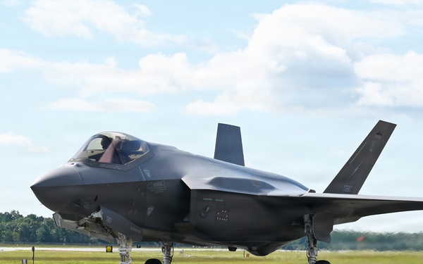 A single-engine stealth multirole fighter aircraft, consistent with an F-35 Lightning II, is taxiing on a paved runway or taxiway. The aircraft is photographed in daylight under partly cloudy skies, with green treelines and grass visible in the background. A pilot is visible in the cockpit, and the landing gear is fully deployed.