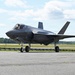 A single-engine stealth multirole fighter aircraft, consistent with an F-35 Lightning II, is taxiing on a paved runway or taxiway. The aircraft is photographed in daylight under partly cloudy skies, with green treelines and grass visible in the background. A pilot is visible in the cockpit, and the landing gear is fully deployed.
