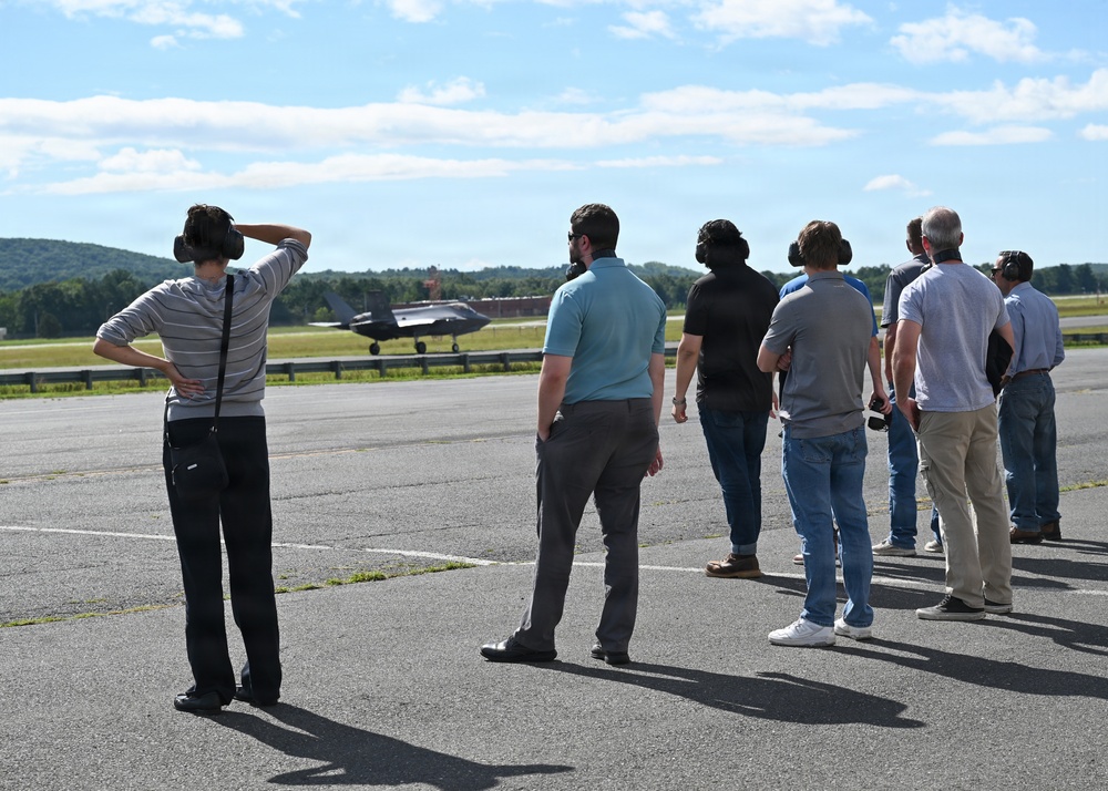 A group of approximately seven to eight civilians stand on an airport tarmac observing a military jet aircraft on the runway in the background. Several individuals are wearing hearing protection ear muffs, suggesting an official airfield visit or demonstration. The setting appears to be a military or joint-use airfield on a clear, sunny day with wooded hills visible in the background.