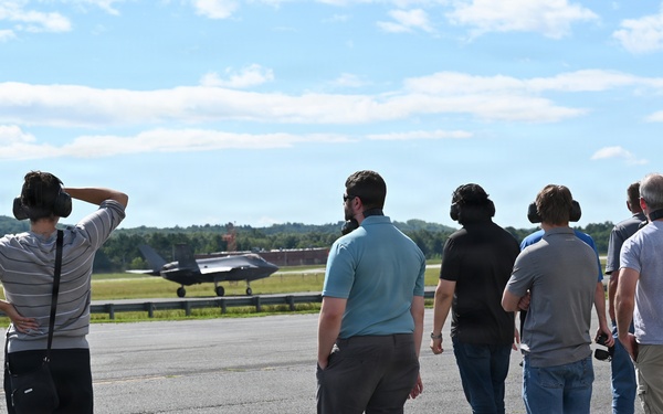 A group of approximately seven to eight civilians stand on an airport tarmac observing a military jet aircraft on the runway in the background. Several individuals are wearing hearing protection ear muffs, suggesting an official airfield visit or demonstration. The setting appears to be a military or joint-use airfield on a clear, sunny day with wooded hills visible in the background.