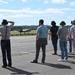 A group of approximately seven to eight civilians stand on an airport tarmac observing a military jet aircraft on the runway in the background. Several individuals are wearing hearing protection ear muffs, suggesting an official airfield visit or demonstration. The setting appears to be a military or joint-use airfield on a clear, sunny day with wooded hills visible in the background.