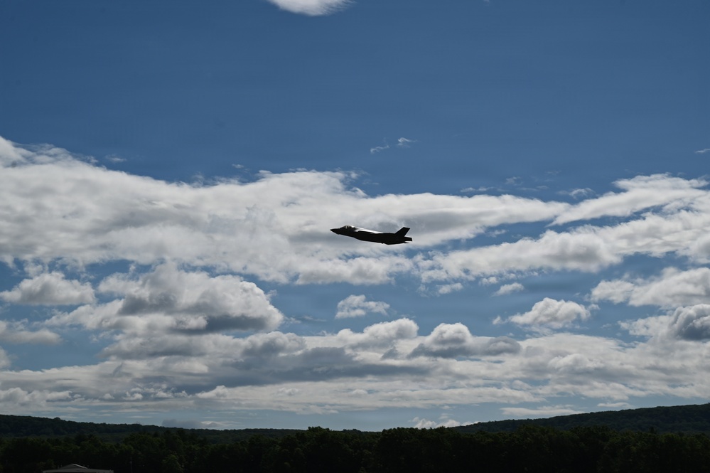 A single military jet aircraft, identifiable by its silhouette as an F-35 Lightning II, is captured in flight against a partly cloudy blue sky. The aircraft appears as a dark silhouette, flying at moderate altitude over a treeline visible at the bottom of the frame. The image was taken from a ground-level perspective looking upward.