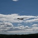 A single military jet aircraft, identifiable by its silhouette as an F-35 Lightning II, is captured in flight against a partly cloudy blue sky. The aircraft appears as a dark silhouette, flying at moderate altitude over a treeline visible at the bottom of the frame. The image was taken from a ground-level perspective looking upward.