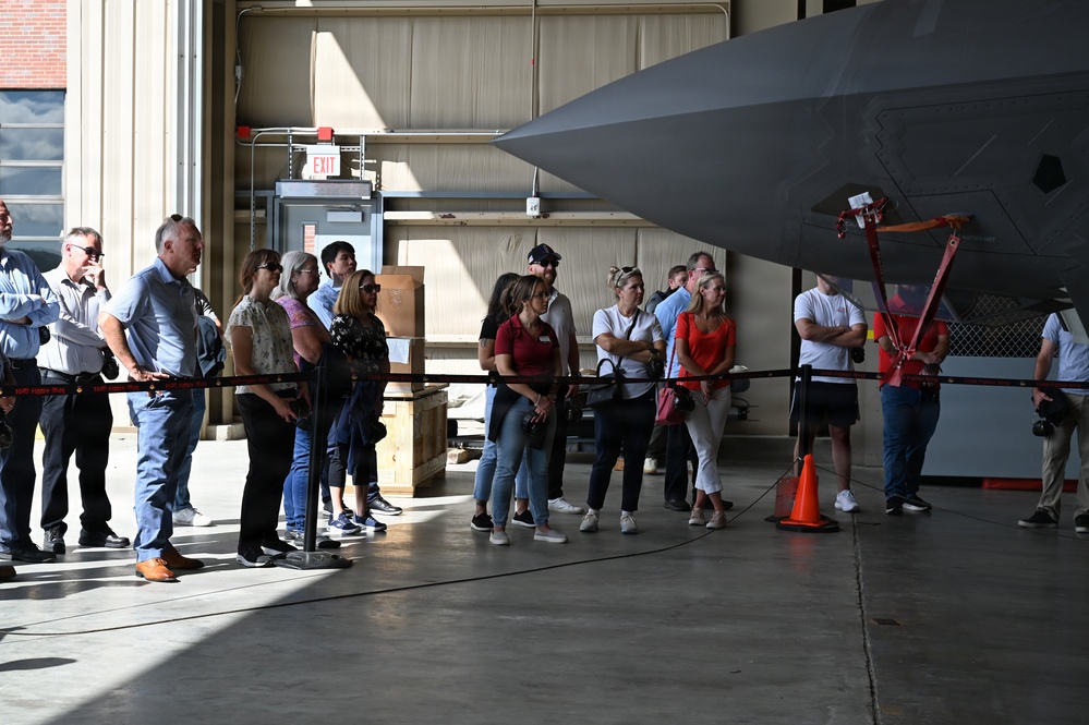 A group of civilians is gathered inside a military aircraft hangar, viewing a large gray stealth aircraft up close. They are standing behind a red rope barrier that separates them from the aircraft, suggesting an organized tour or public affairs event. The setting appears to be an active military installation based on the hangar infrastructure and safety equipment visible.