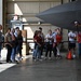 A group of civilians is gathered inside a military aircraft hangar, viewing a large gray stealth aircraft up close. They are standing behind a red rope barrier that separates them from the aircraft, suggesting an organized tour or public affairs event. The setting appears to be an active military installation based on the hangar infrastructure and safety equipment visible.