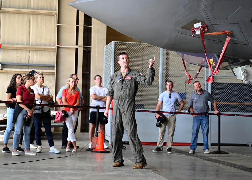 A military pilot in an olive-green flight suit is conducting a briefing or tour for a group of civilians inside an aircraft hangar. He is pointing upward toward a large military aircraft, likely explaining features of the aircraft to the assembled visitors. The group stands behind a rope barrier, suggesting this is an organized public or community relations event.