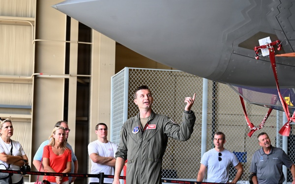 A military pilot in an olive-green flight suit is conducting a briefing or tour for a group of civilians inside an aircraft hangar. He is pointing upward toward a large military aircraft, likely explaining features of the aircraft to the assembled visitors. The group stands behind a rope barrier, suggesting this is an organized public or community relations event.