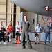 A military pilot in an olive-green flight suit is conducting a briefing or tour for a group of civilians inside an aircraft hangar. He is pointing upward toward a large military aircraft, likely explaining features of the aircraft to the assembled visitors. The group stands behind a rope barrier, suggesting this is an organized public or community relations event.