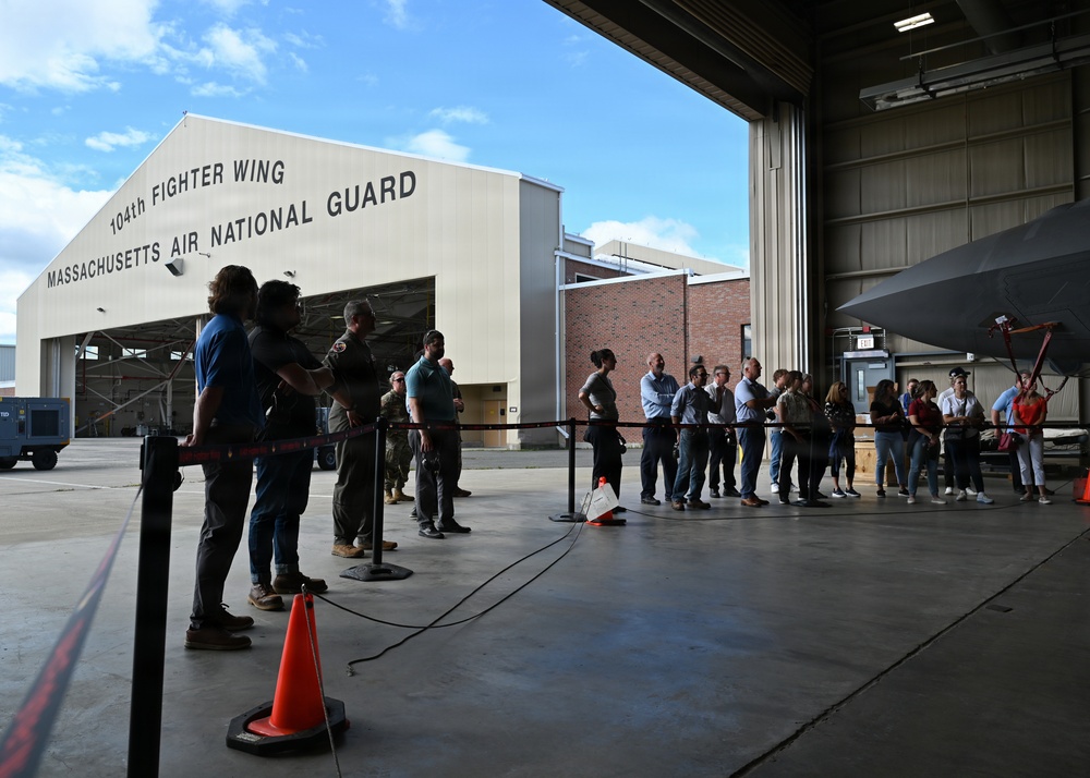 A group of civilians and military personnel are gathered inside a hangar at the 104th Fighter Wing, Massachusetts Air National Guard base. The visitors appear to be on an organized tour or briefing event, standing behind rope barriers near a military aircraft. The scene takes place on a clear, partly cloudy day with the unit's main hangar building visible in the background.