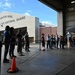 A group of civilians and military personnel are gathered inside a hangar at the 104th Fighter Wing, Massachusetts Air National Guard base. The visitors appear to be on an organized tour or briefing event, standing behind rope barriers near a military aircraft. The scene takes place on a clear, partly cloudy day with the unit's main hangar building visible in the background.