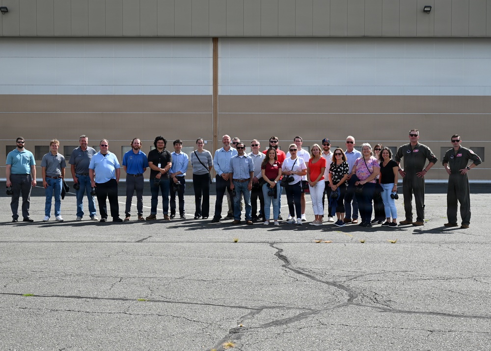 A large group of approximately 25-30 people are posed for a group photograph in front of a large industrial or military hangar building. Two individuals on the far right appear to be wearing military flight suits, suggesting this may be an official visit or tour of a military aviation facility. The group consists of men and women in civilian attire, standing on a paved tarmac or apron area.