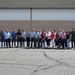 A large group of approximately 25-30 people are posed for a group photograph in front of a large industrial or military hangar building. Two individuals on the far right appear to be wearing military flight suits, suggesting this may be an official visit or tour of a military aviation facility. The group consists of men and women in civilian attire, standing on a paved tarmac or apron area.