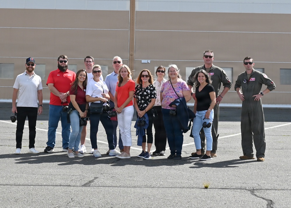 A group of approximately 13 people pose for a group photo on what appears to be a military airfield tarmac in front of a large hangar building. Two individuals on the right side of the group are wearing olive drab military flight suits, suggesting this may be a civilian visit or tour of a military installation. Several members of the group are carrying cameras and camera bags.