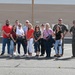A group of approximately 13 people pose for a group photo on what appears to be a military airfield tarmac in front of a large hangar building. Two individuals on the right side of the group are wearing olive drab military flight suits, suggesting this may be a civilian visit or tour of a military installation. Several members of the group are carrying cameras and camera bags.