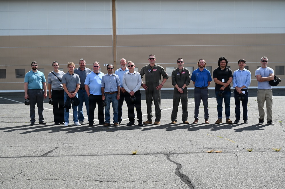 A group of approximately 14 men are posing for a group photograph outdoors in front of a large industrial or hangar-style building. Two individuals in the center are wearing military flight suits, suggesting this may be a military base or aviation facility visit. The group appears to consist of a mix of military personnel and civilian visitors or contractors.