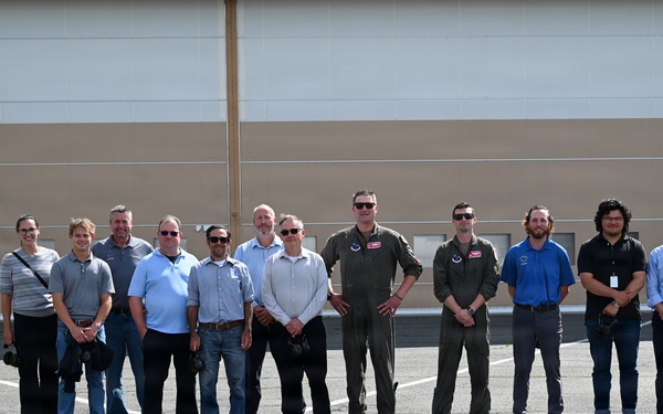 A group of approximately 14 men are posing for a group photograph outdoors in front of a large industrial or hangar-style building. Two individuals in the center are wearing military flight suits, suggesting this may be a military base or aviation facility visit. The group appears to consist of a mix of military personnel and civilian visitors or contractors.