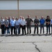 A group of approximately 14 men are posing for a group photograph outdoors in front of a large industrial or hangar-style building. Two individuals in the center are wearing military flight suits, suggesting this may be a military base or aviation facility visit. The group appears to consist of a mix of military personnel and civilian visitors or contractors.