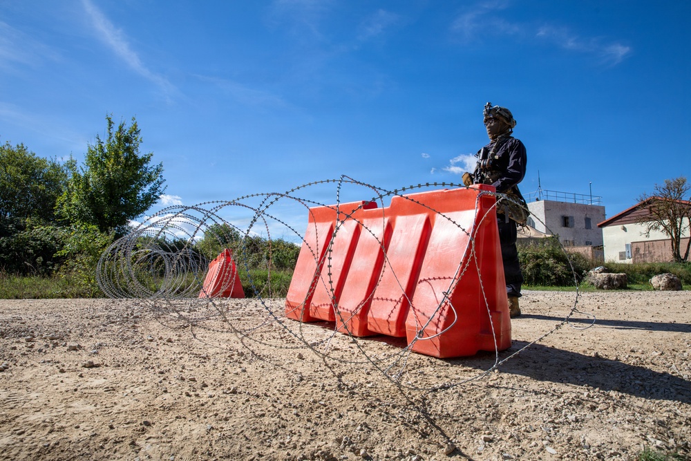 A uniformed armed individual stands behind a row of bright red concrete security barriers reinforced with concertina/razor wire coils on a dirt road. The scene depicts a security checkpoint or defensive position in what appears to be a semi-rural area with buildings and vegetation visible in the background. The setup suggests a deliberate access-control or force-protection measure.