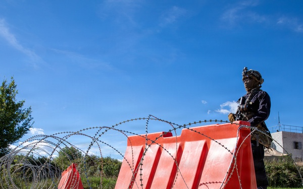 A uniformed armed individual stands behind a row of bright red concrete security barriers reinforced with concertina/razor wire coils on a dirt road. The scene depicts a security checkpoint or defensive position in what appears to be a semi-rural area with buildings and vegetation visible in the background. The setup suggests a deliberate access-control or force-protection measure.