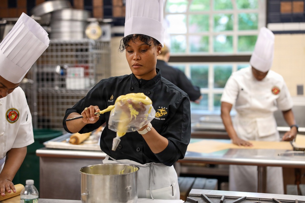 A professional kitchen scene featuring multiple chefs in uniform working at their stations. The central figure, wearing a black chef's coat with a U.S. Marine Corps emblem and the text 'Marine Aide,' is filling a piping bag with what appears to be a yellow batter or cream. The setting appears to be a military culinary training or official cooking facility.