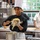 A professional kitchen scene featuring multiple chefs in uniform working at their stations. The central figure, wearing a black chef's coat with a U.S. Marine Corps emblem and the text 'Marine Aide,' is filling a piping bag with what appears to be a yellow batter or cream. The setting appears to be a military culinary training or official cooking facility.