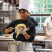 A professional kitchen scene featuring multiple chefs in uniform working at their stations. The central figure, wearing a black chef's coat with a U.S. Marine Corps emblem and the text 'Marine Aide,' is filling a piping bag with what appears to be a yellow batter or cream. The setting appears to be a military culinary training or official cooking facility.