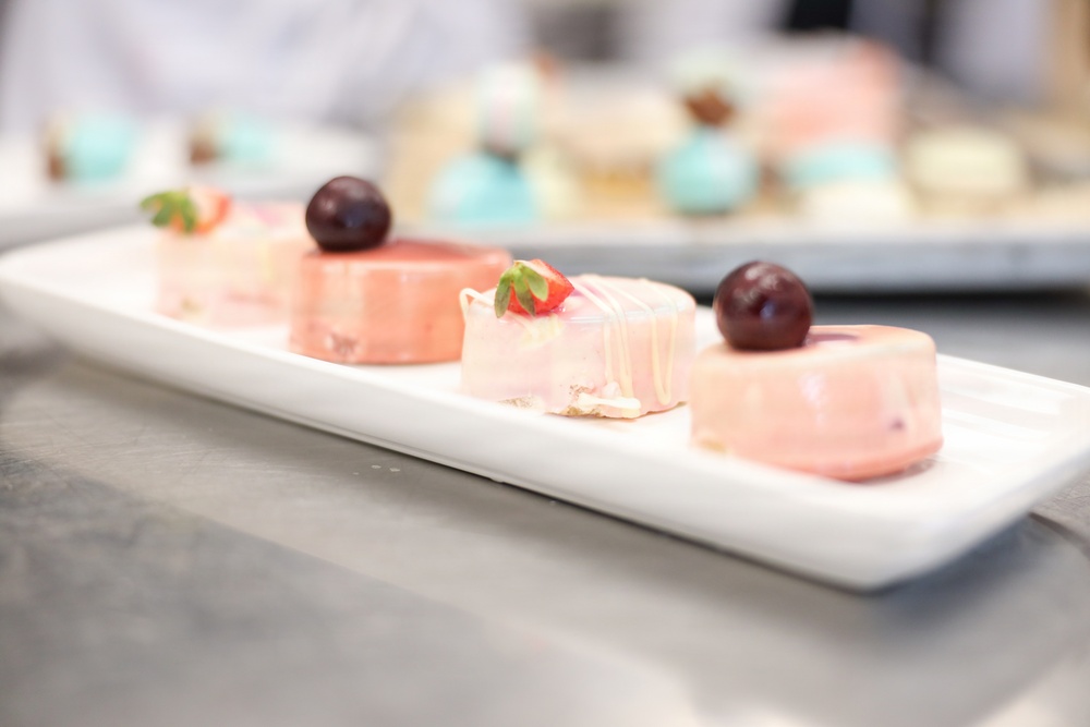 A white rectangular serving platter holds four small pink desserts, likely petit fours or mousse cakes, garnished with dark cherries and a strawberry slice. The desserts have a smooth pink glaze finish with one appearing to have a marbled white-and-pink pattern. In the blurred background, additional desserts on a separate tray are visible, suggesting a professional pastry or catering setting.