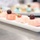 A white rectangular serving platter holds four small pink desserts, likely petit fours or mousse cakes, garnished with dark cherries and a strawberry slice. The desserts have a smooth pink glaze finish with one appearing to have a marbled white-and-pink pattern. In the blurred background, additional desserts on a separate tray are visible, suggesting a professional pastry or catering setting.