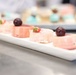 A white rectangular serving platter holds four small pink desserts, likely petit fours or mousse cakes, garnished with dark cherries and a strawberry slice. The desserts have a smooth pink glaze finish with one appearing to have a marbled white-and-pink pattern. In the blurred background, additional desserts on a separate tray are visible, suggesting a professional pastry or catering setting.