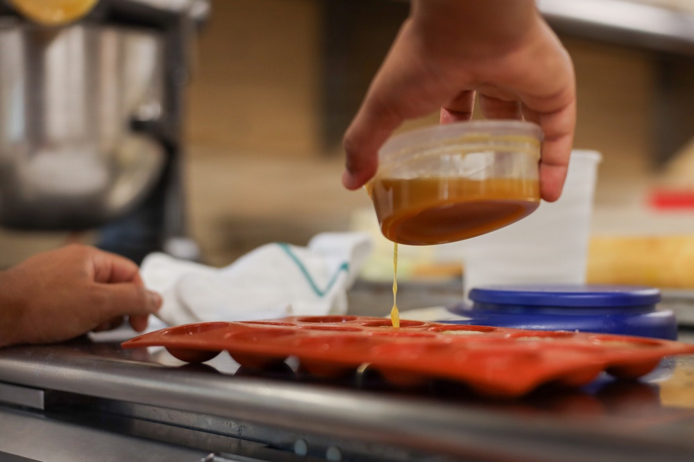 A person is pouring a golden-brown liquid, likely caramel or honey, from a small clear plastic container into a red silicone mold tray. The scene takes place in what appears to be a commercial or professional kitchen setting. A second person's hand is visible in the background, steadying the tray.