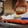 A person is pouring a golden-brown liquid, likely caramel or honey, from a small clear plastic container into a red silicone mold tray. The scene takes place in what appears to be a commercial or professional kitchen setting. A second person's hand is visible in the background, steadying the tray.