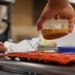 A person is pouring a golden-brown liquid, likely caramel or honey, from a small clear plastic container into a red silicone mold tray. The scene takes place in what appears to be a commercial or professional kitchen setting. A second person's hand is visible in the background, steadying the tray.