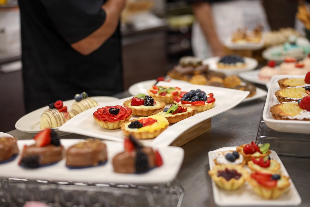 The image shows a professional dessert display featuring an assortment of fruit tarts, chocolate pastries, and decorated confections arranged on white serving platters. Multiple trays are spread across what appears to be a stainless steel buffet or catering counter. At least one person wearing a black chef's uniform is visible in the background, suggesting a professional kitchen or catering event setting.