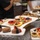 The image shows a professional dessert display featuring an assortment of fruit tarts, chocolate pastries, and decorated confections arranged on white serving platters. Multiple trays are spread across what appears to be a stainless steel buffet or catering counter. At least one person wearing a black chef's uniform is visible in the background, suggesting a professional kitchen or catering event setting.