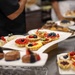 The image shows a professional dessert display featuring an assortment of fruit tarts, chocolate pastries, and decorated confections arranged on white serving platters. Multiple trays are spread across what appears to be a stainless steel buffet or catering counter. At least one person wearing a black chef's uniform is visible in the background, suggesting a professional kitchen or catering event setting.