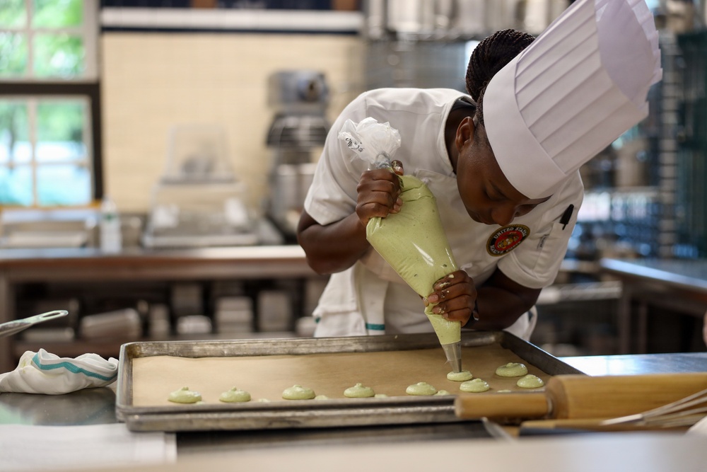 A pastry chef in a white uniform and toque is carefully piping a light green batter or cream onto a parchment-lined baking sheet, likely making macarons or similar confections. The setting is a professional kitchen with commercial equipment visible in the background. The chef is leaning in closely, demonstrating precision and focus in the task.