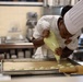 A pastry chef in a white uniform and toque is carefully piping a light green batter or cream onto a parchment-lined baking sheet, likely making macarons or similar confections. The setting is a professional kitchen with commercial equipment visible in the background. The chef is leaning in closely, demonstrating precision and focus in the task.