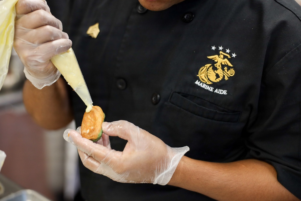 A person wearing a black 'Marine Aide' uniform with the U.S. Marine Corps Eagle, Globe, and Anchor emblem is preparing food in a kitchen setting. They are using a piping bag to apply a yellow sauce or cream onto a small food item, likely a canapé or appetizer. The individual is wearing clear food-safe gloves, indicating professional food handling standards.