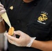 A person wearing a black 'Marine Aide' uniform with the U.S. Marine Corps Eagle, Globe, and Anchor emblem is preparing food in a kitchen setting. They are using a piping bag to apply a yellow sauce or cream onto a small food item, likely a canapé or appetizer. The individual is wearing clear food-safe gloves, indicating professional food handling standards.