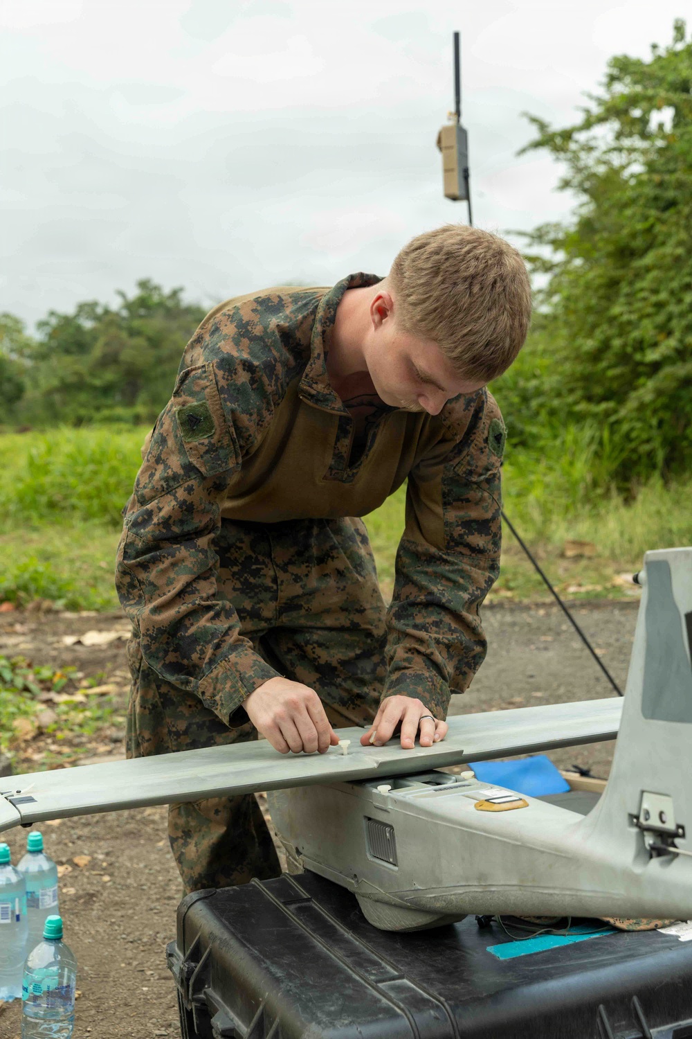 A military service member in MARPAT (Marine pattern) camouflage uniform is working on what appears to be a small fixed-wing UAV (unmanned aerial vehicle), likely a RQ-11 Raven or similar hand-launched drone. The individual is assembling or inspecting the aircraft's wing components on top of a hard-shell Pelican-style transport case. The setting appears to be an outdoor field environment with lush green vegetation in the background.